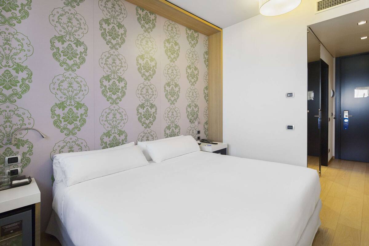A hotel room with a large bed, a bedside table with a lamp, and a wall with a patterned wallpaper.