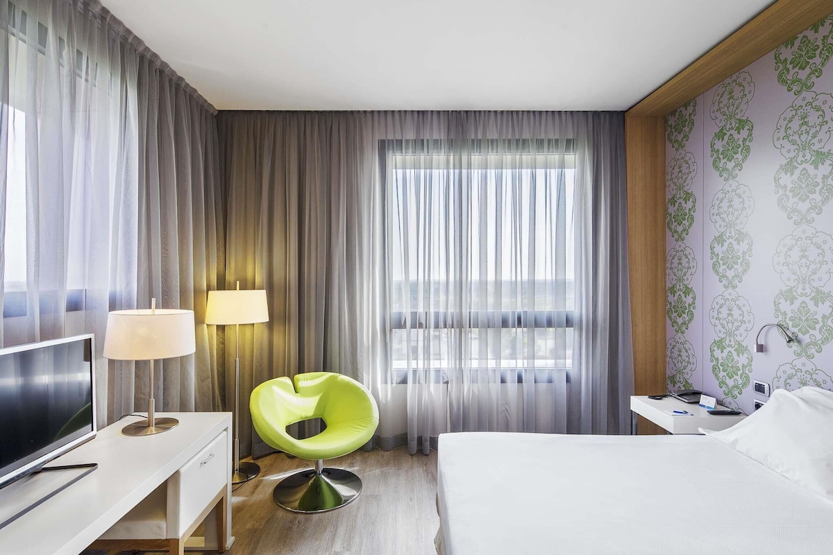 A hotel room with a bed, a TV, a desk with a lamp, a green chair, and a window with curtains.