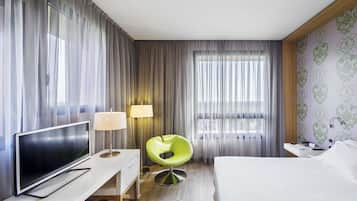 Une chambre d’hôtel avec un lit, une télévision, un bureau avec une lampe, une chaise verte et une fenêtre avec des rideaux.