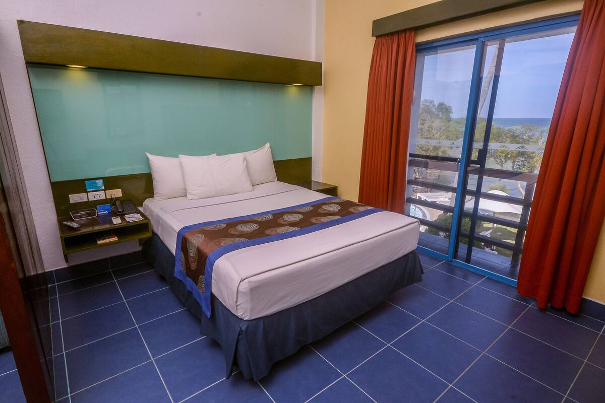 A hotel room with a bed, a nightstand, a window with curtains, and a view of the outside.