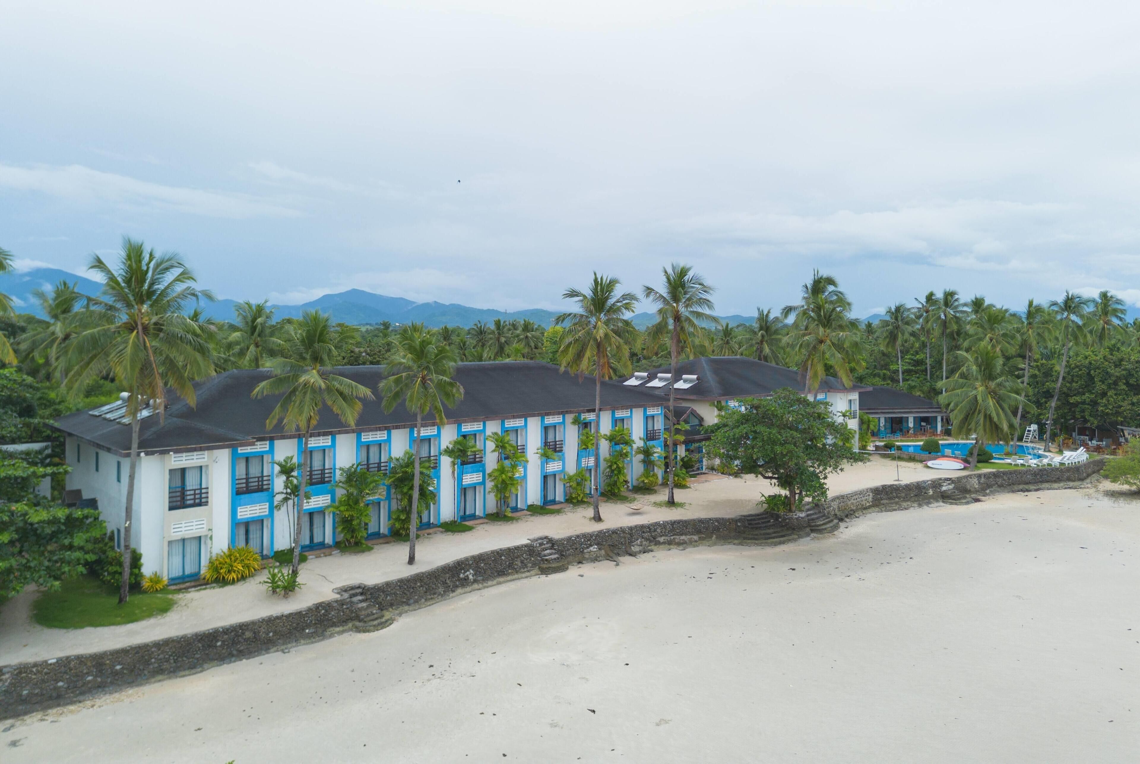 Un hotel frente a la playa con varias habitaciones, piscina y palmeras.