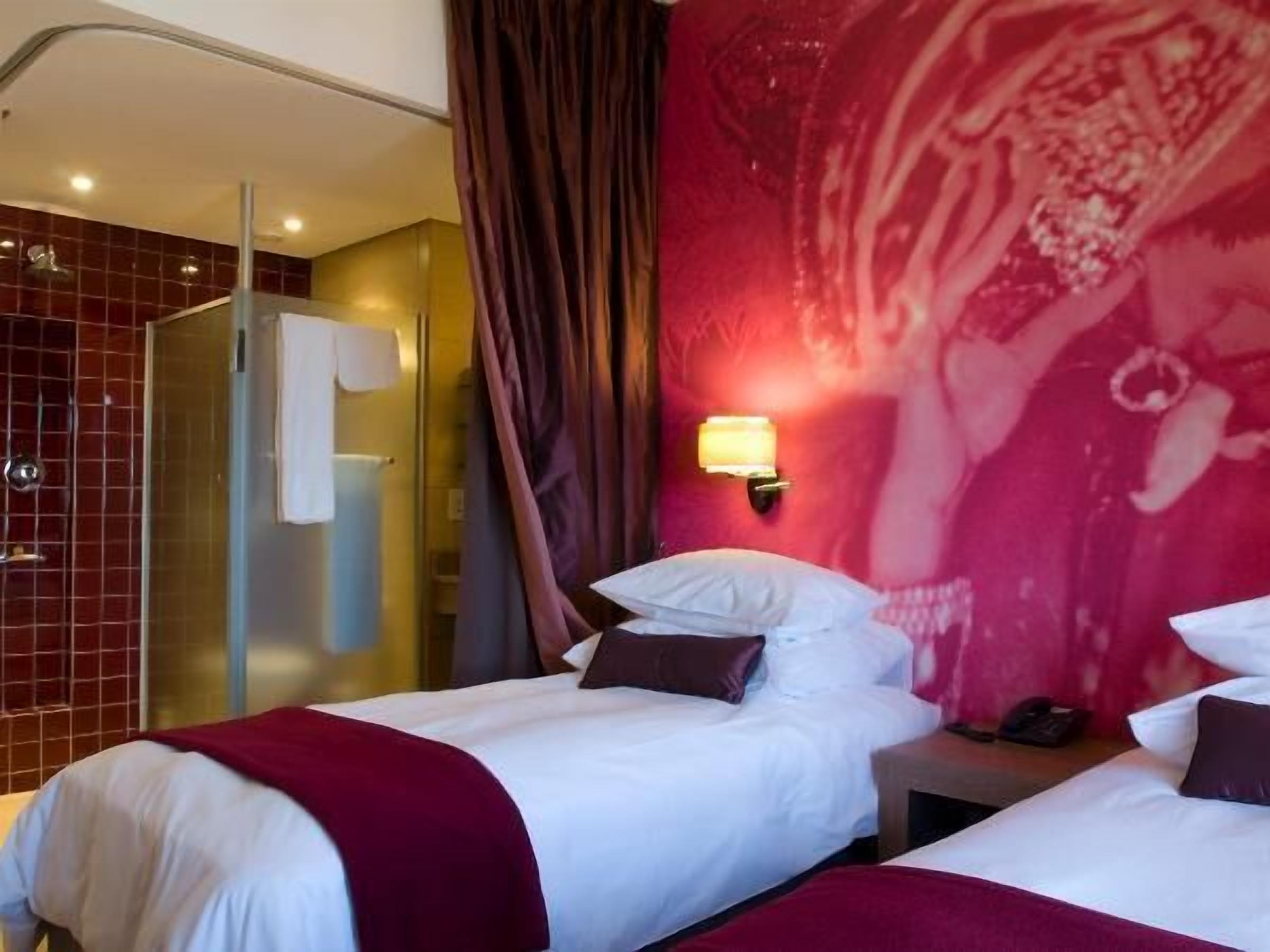 Camera d'albergo con due letti, cabina doccia in vetro, parete con piastrelle rosse e luce a parete.