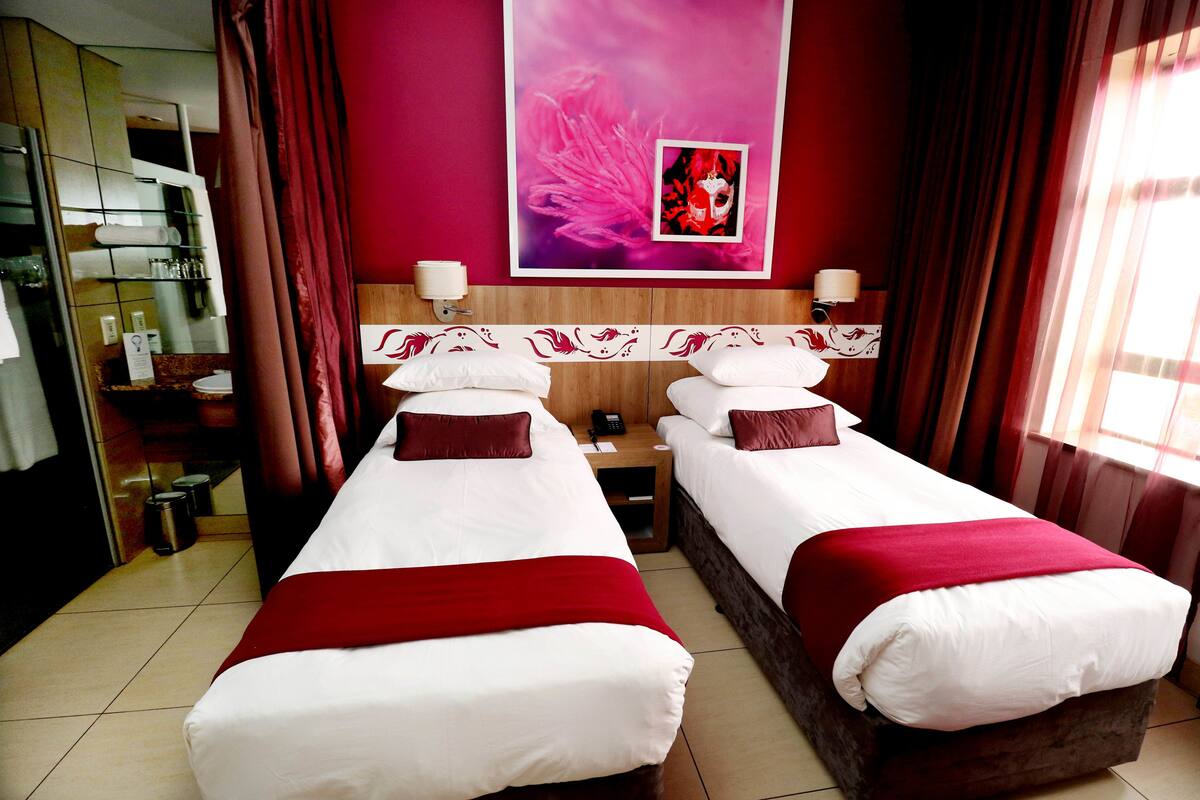 A hotel room with two beds, a large mirror, a red and white wall art piece, and a window with curtains.