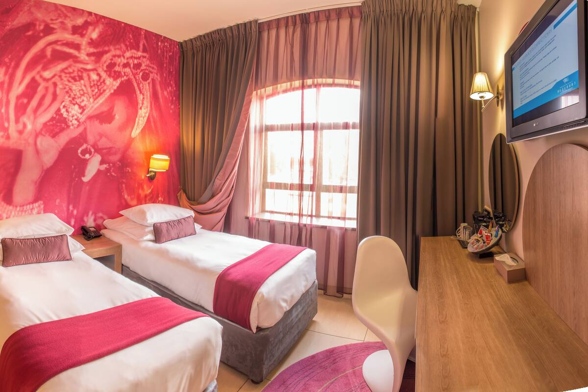 A hotel room with two beds, a shower, a vanity with a mirror, a TV, and a large window with curtains.