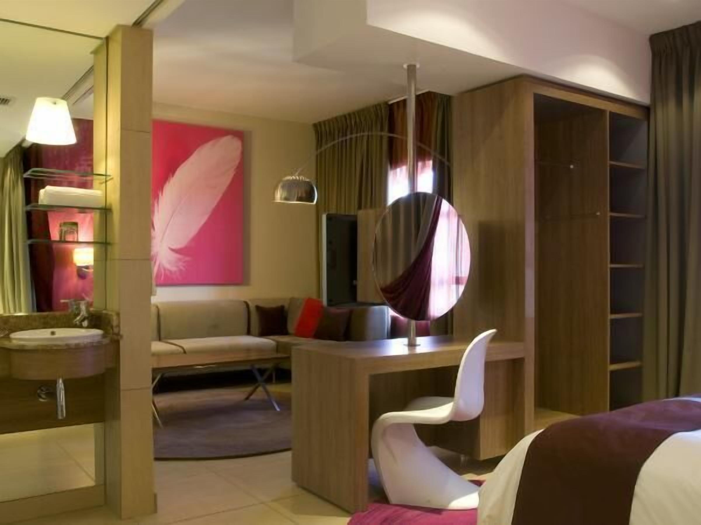 A modern hotel room with a bed, a desk, a chair, a round mirror, a wardrobe, and a bathroom with a sink and mirror.