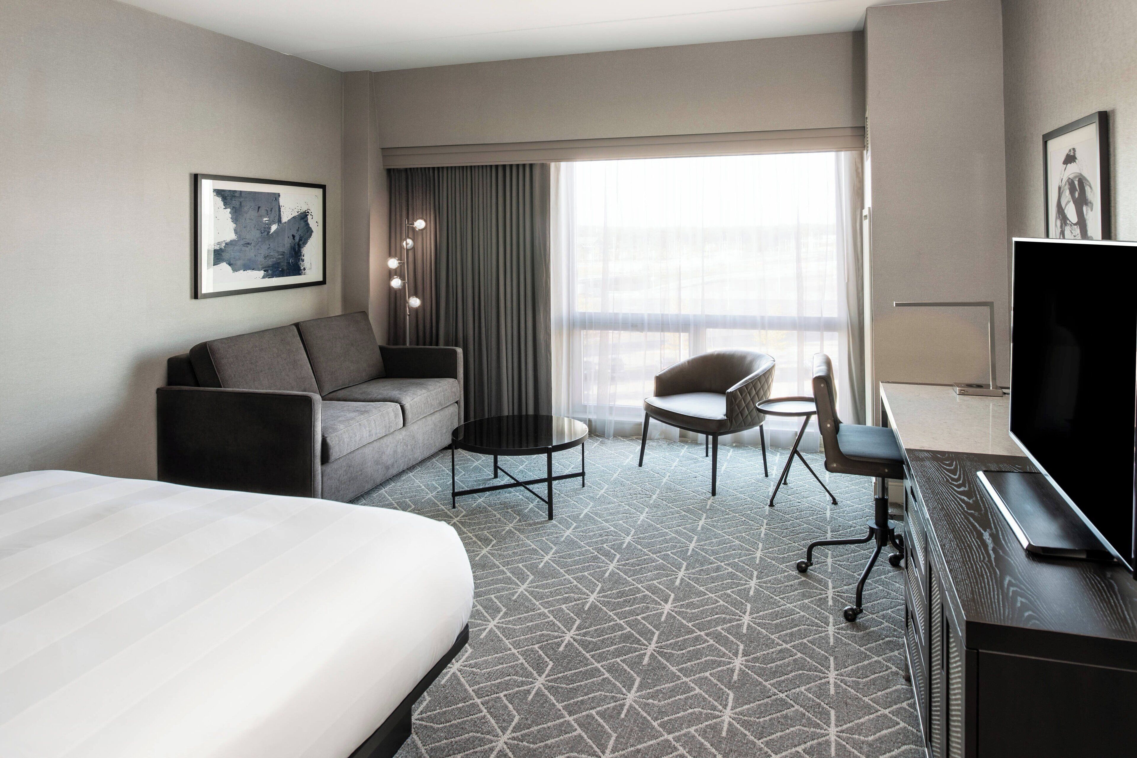 A hotel room with a large bed, a sofa, a desk with a TV, a chair, a small table, and a large window with curtains.