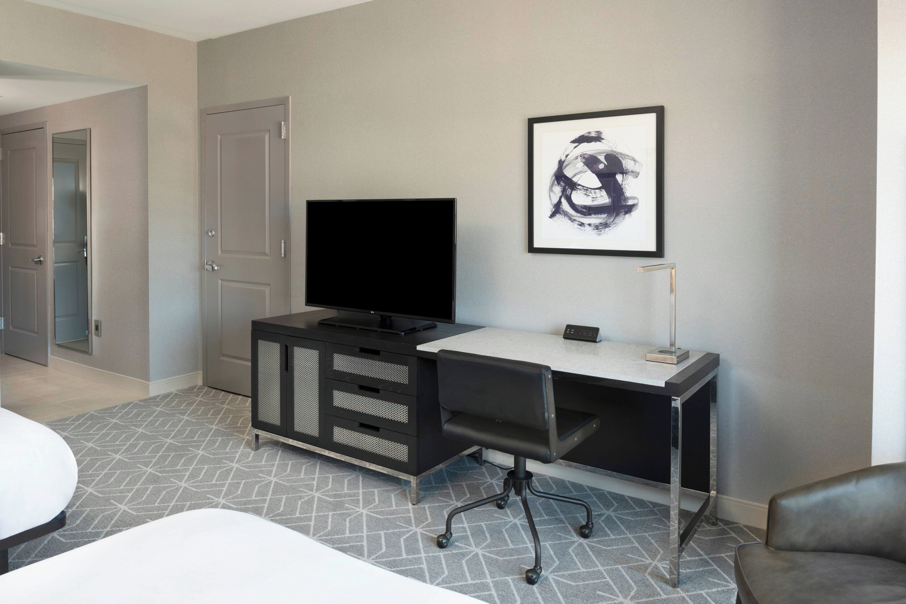 A hotel room with a bed, a desk with a computer, a TV, and a framed artwork on the wall.
