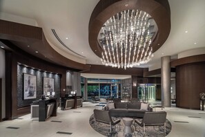 A modern lobby with a central chandelier, a reception desk, seating area, and large windows offering a view of the outside. - Renaissance by Marriott Boston Patriot Place Hotel (Foxborough)