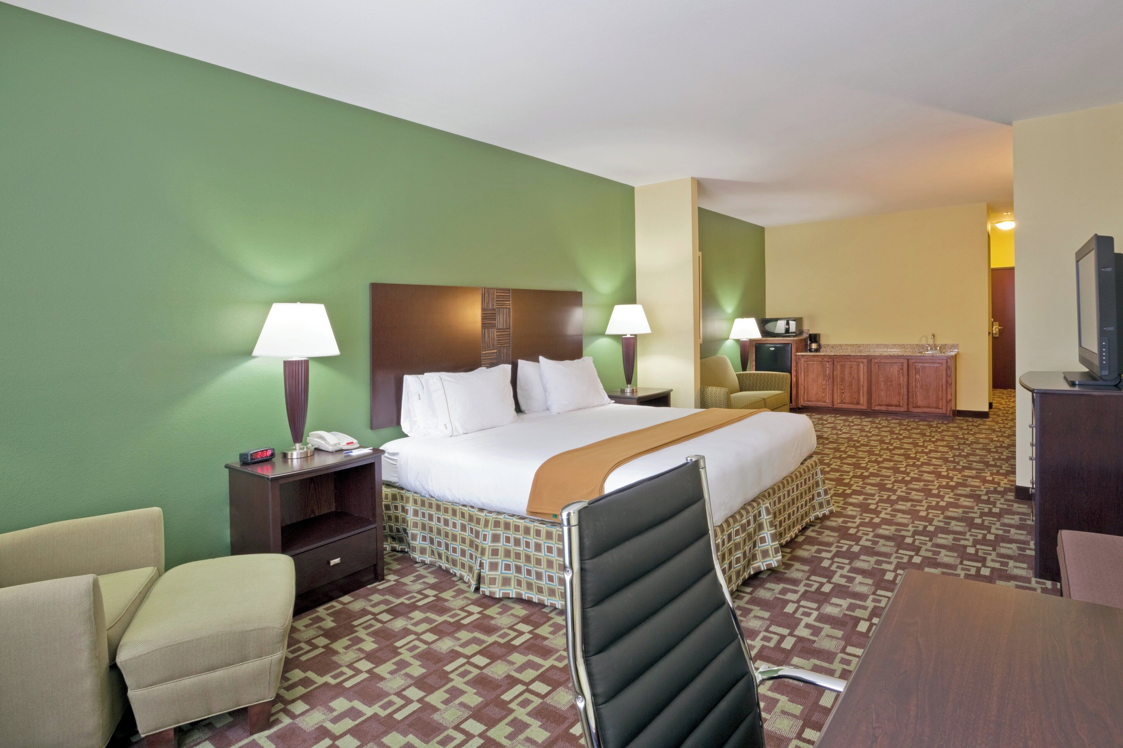 Habitación de hotel con cama, dos mesitas de noche, una silla, escritorio, televisor y kitchenette.