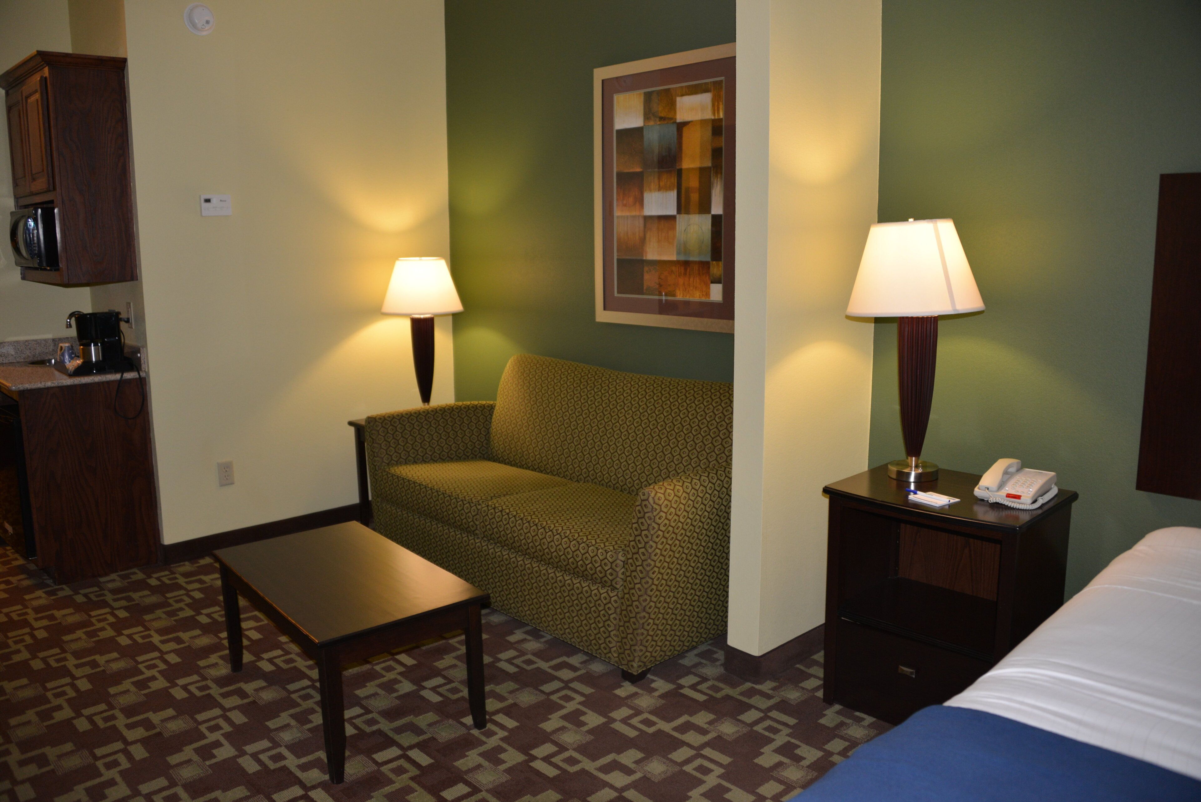 A hotel room with a green patterned sofa, a small table, a lamp, a bedside table with a phone, and a microwave in the corner.