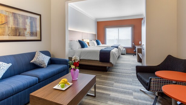 A hotel room with a blue sofa, a wooden coffee table with fruit, a bed with white bedding, and a view of the city through large windows.
