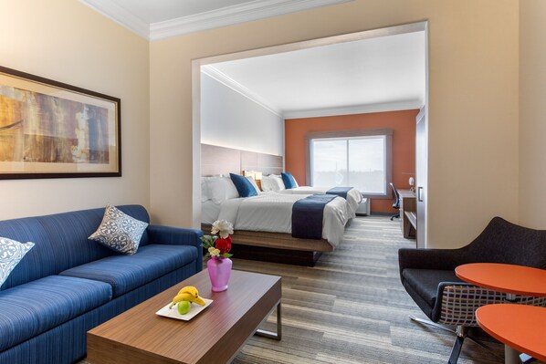 A hotel room with a blue sofa, a wooden coffee table with fruit, a bed with white bedding, and a view of the city through large windows.