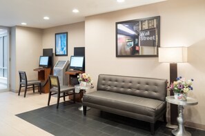 A hotel lobby with a brown leather sofa, two chairs, a small round table with a lamp, a reception desk with a computer, and a framed picture of Wall Street. - Holiday Inn Express New York City- Wall Street by IHG (New York)