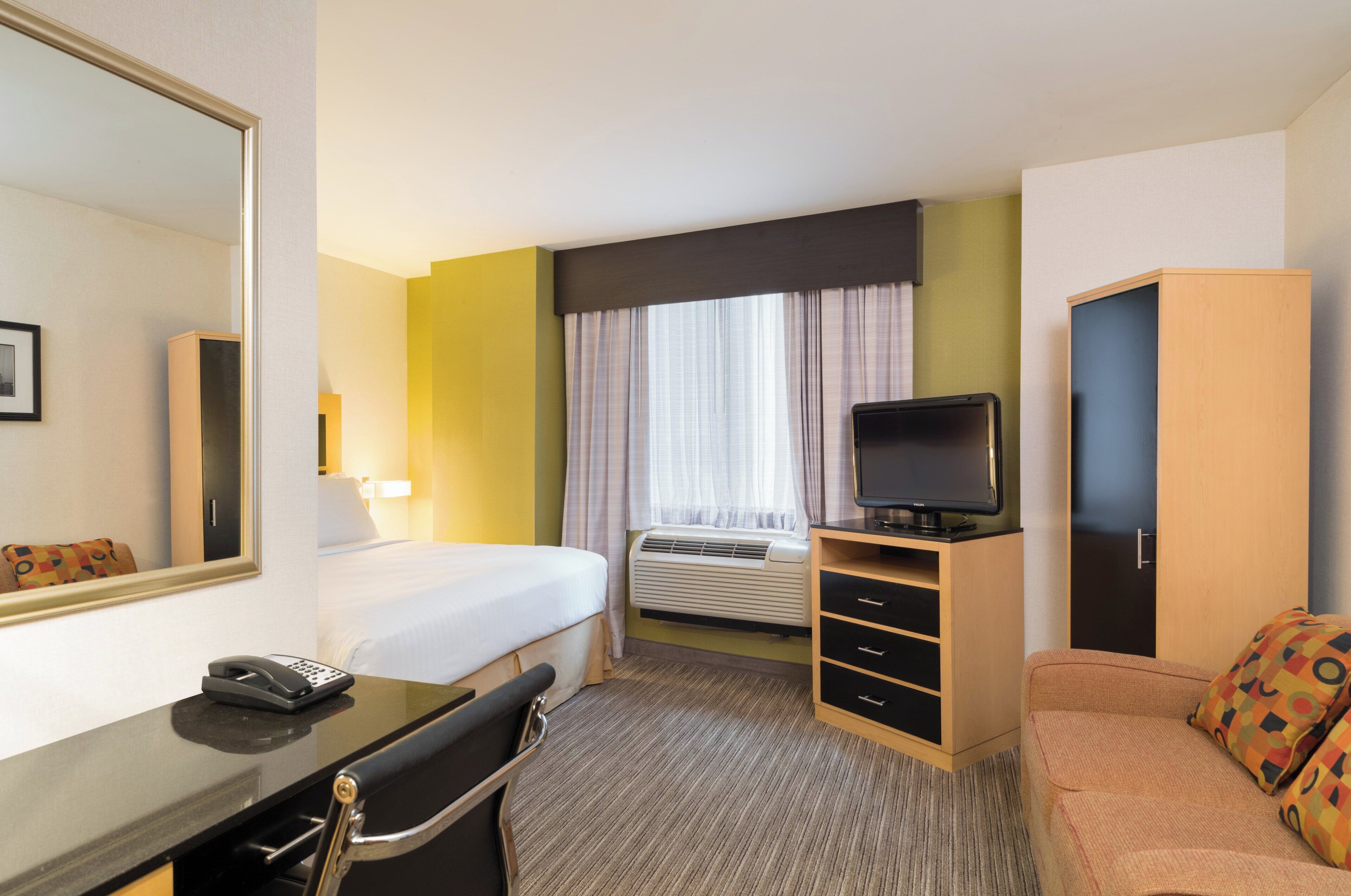 A hotel room with a bed, a desk with a telephone, a television, an air conditioning unit, and a sofa.