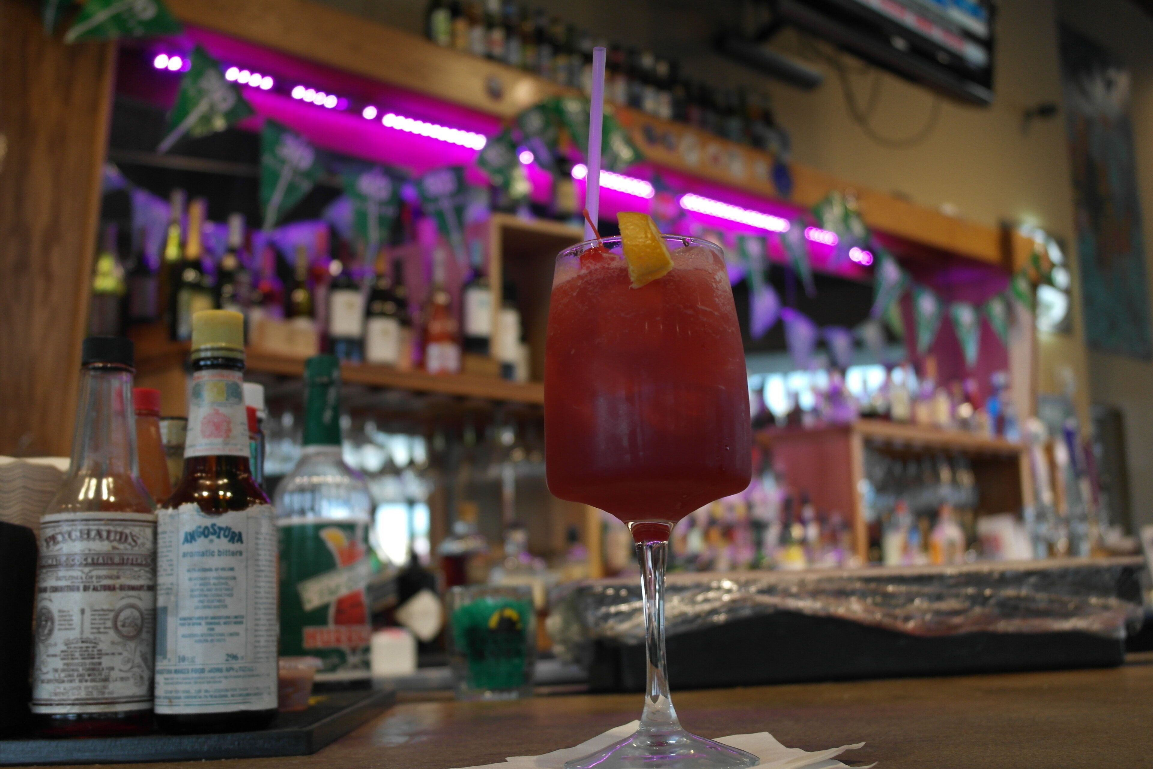 Un bar con una copa de bebida roja, botellas y estanterías al fondo.