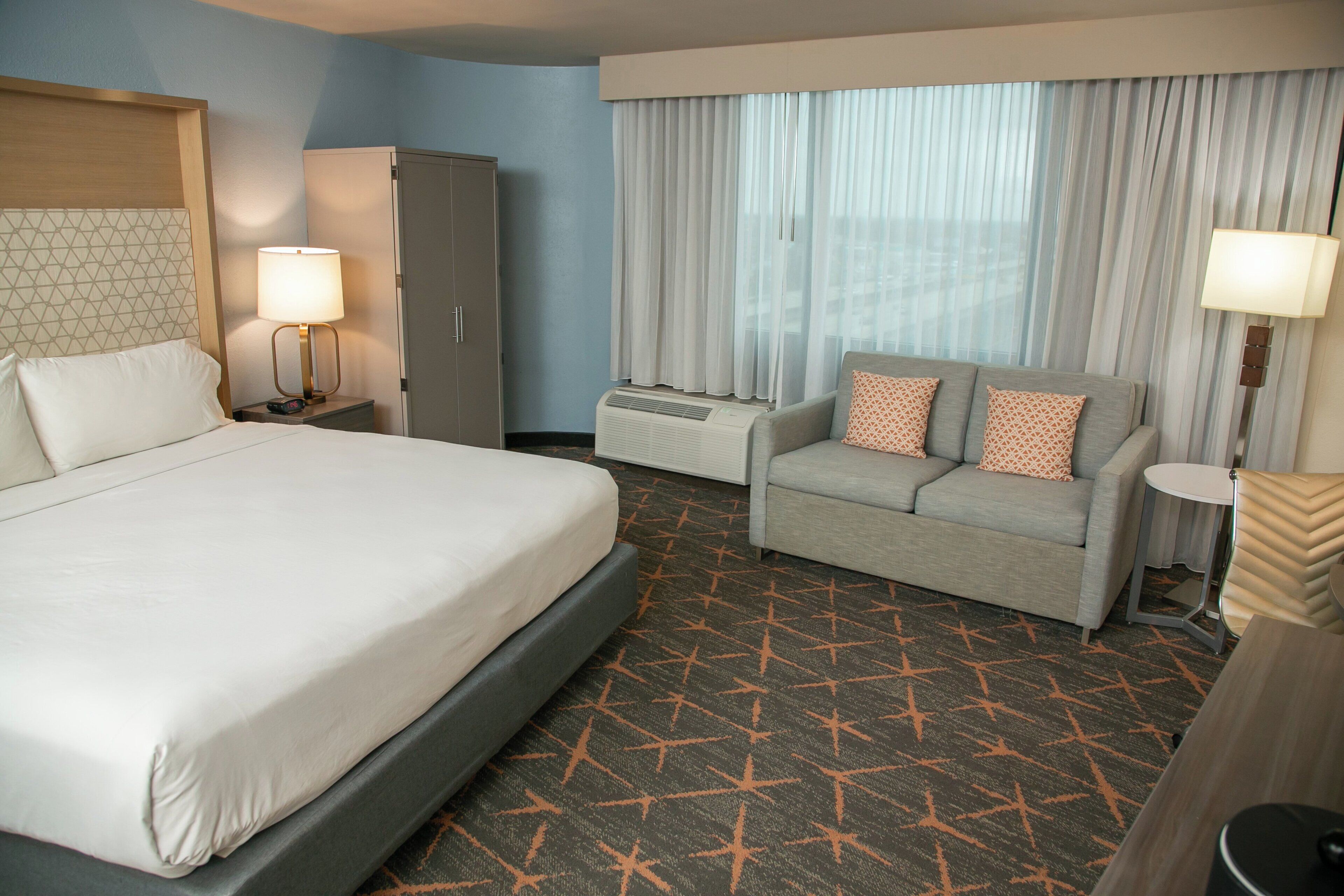 A hotel room with a large bed, a sofa, a desk, a wardrobe, and a window with curtains.