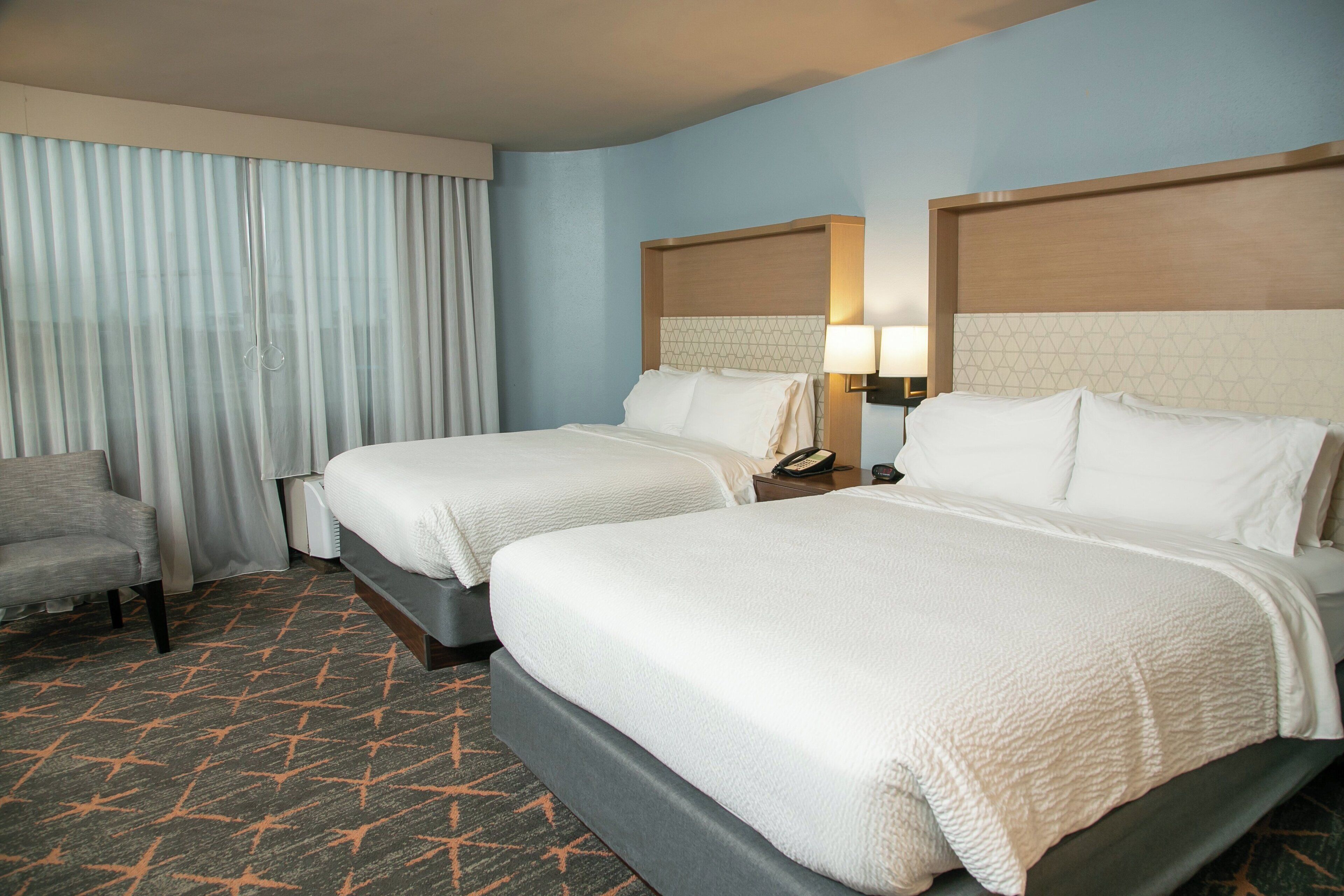 A hotel room with two beds, a chair, a nightstand with a lamp, and a window with curtains.
