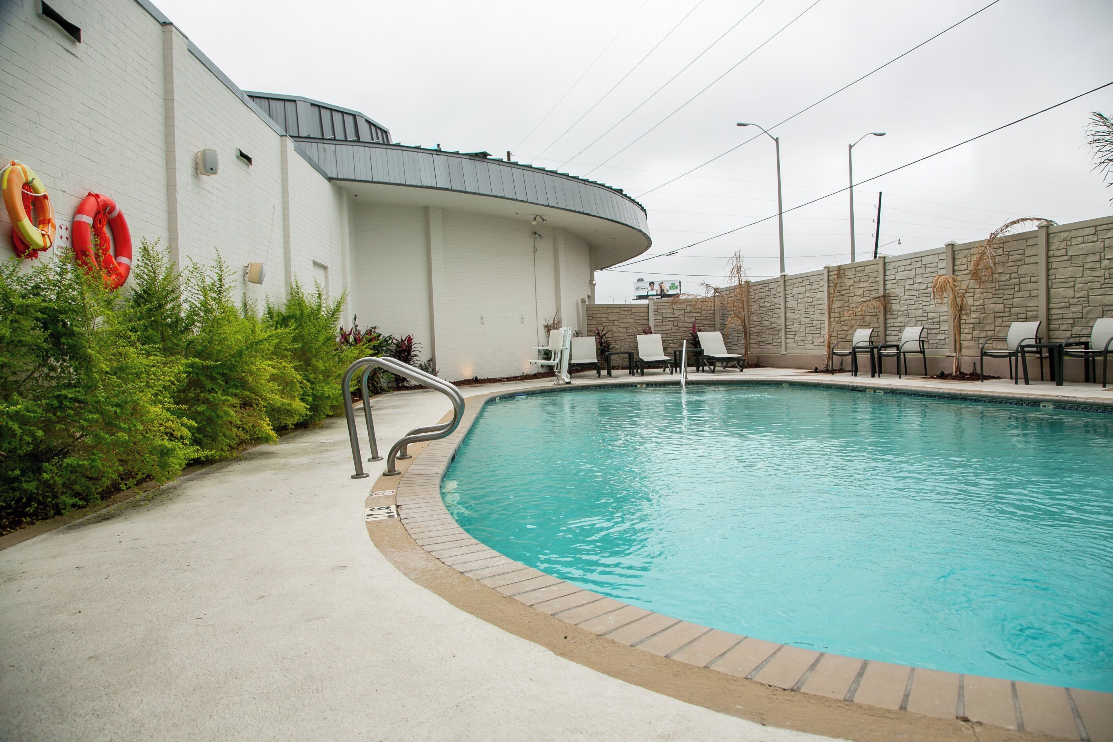 A swimming pool with a curved edge, adjacent to a white building with a curved roof, surrounded by lounge chairs and a stone wall.