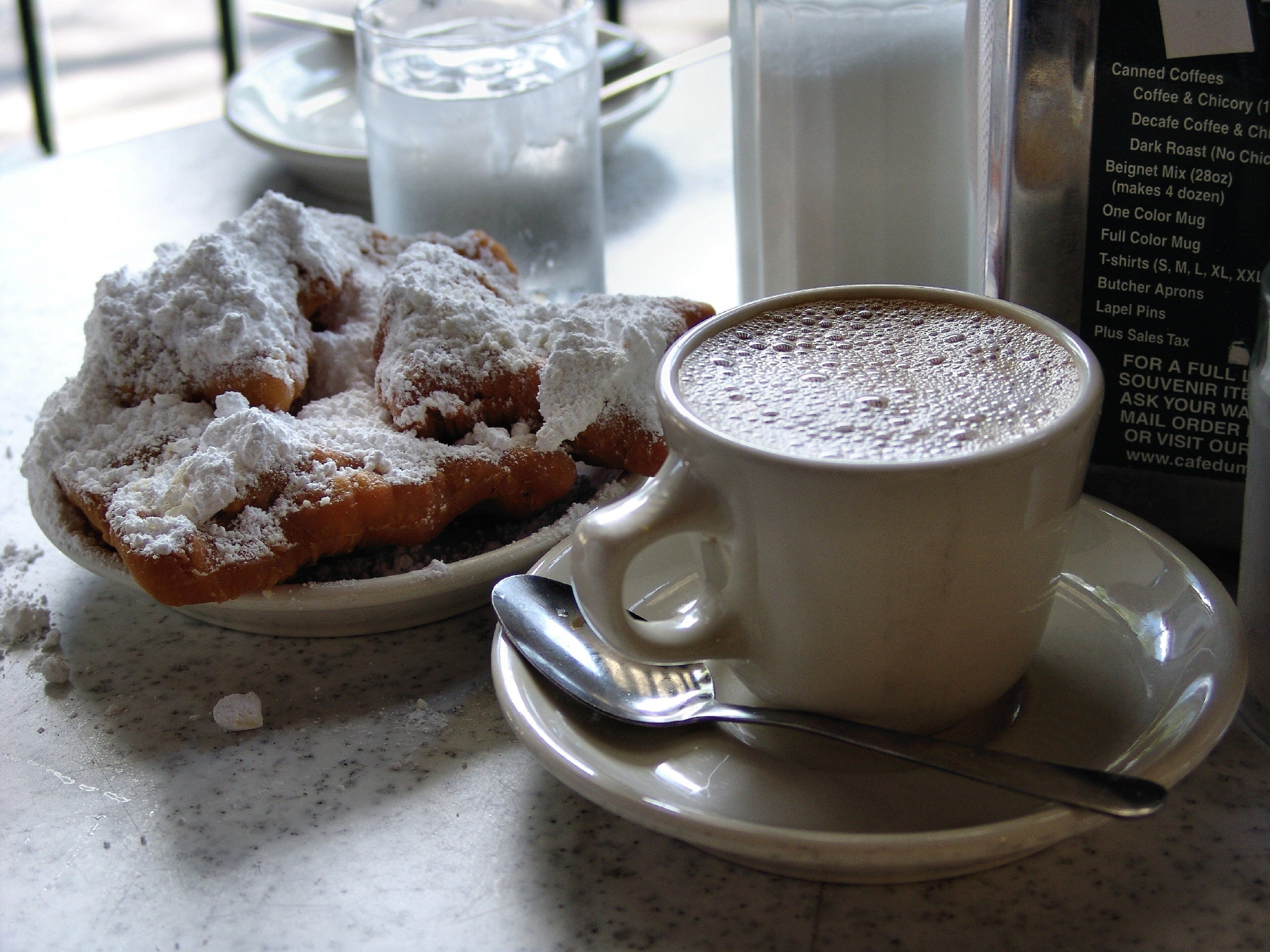 A cup of coffee with a spoon on a saucer, a plate of powdered sugar sprinkled pastries, and a menu on a table.