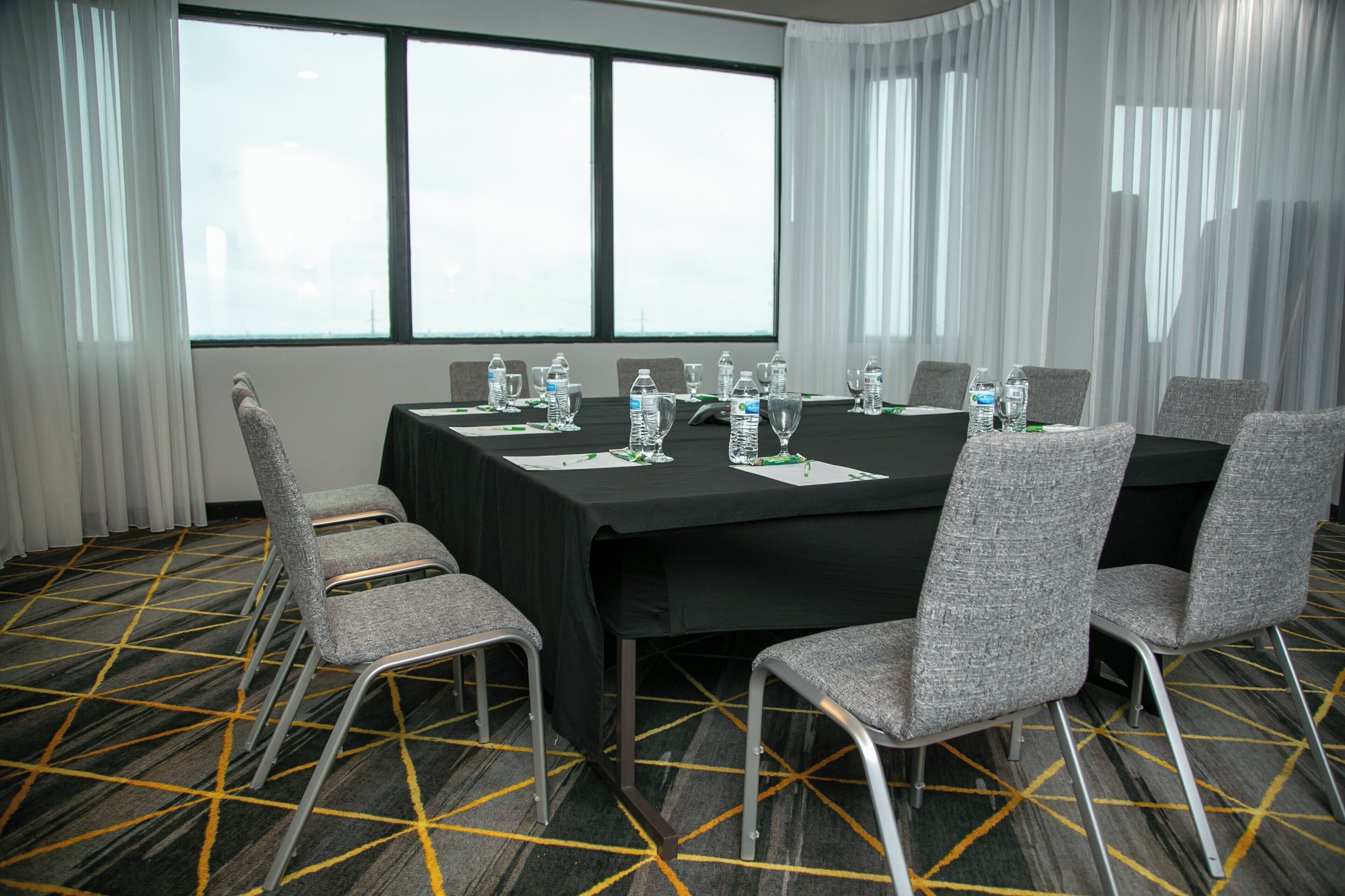 Un salón de conferencias con una mesa negra, sillas grises, botellas de agua y una alfombra estampada.