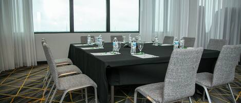 Un salón de conferencias con una mesa negra, sillas grises, botellas de agua y una alfombra estampada.