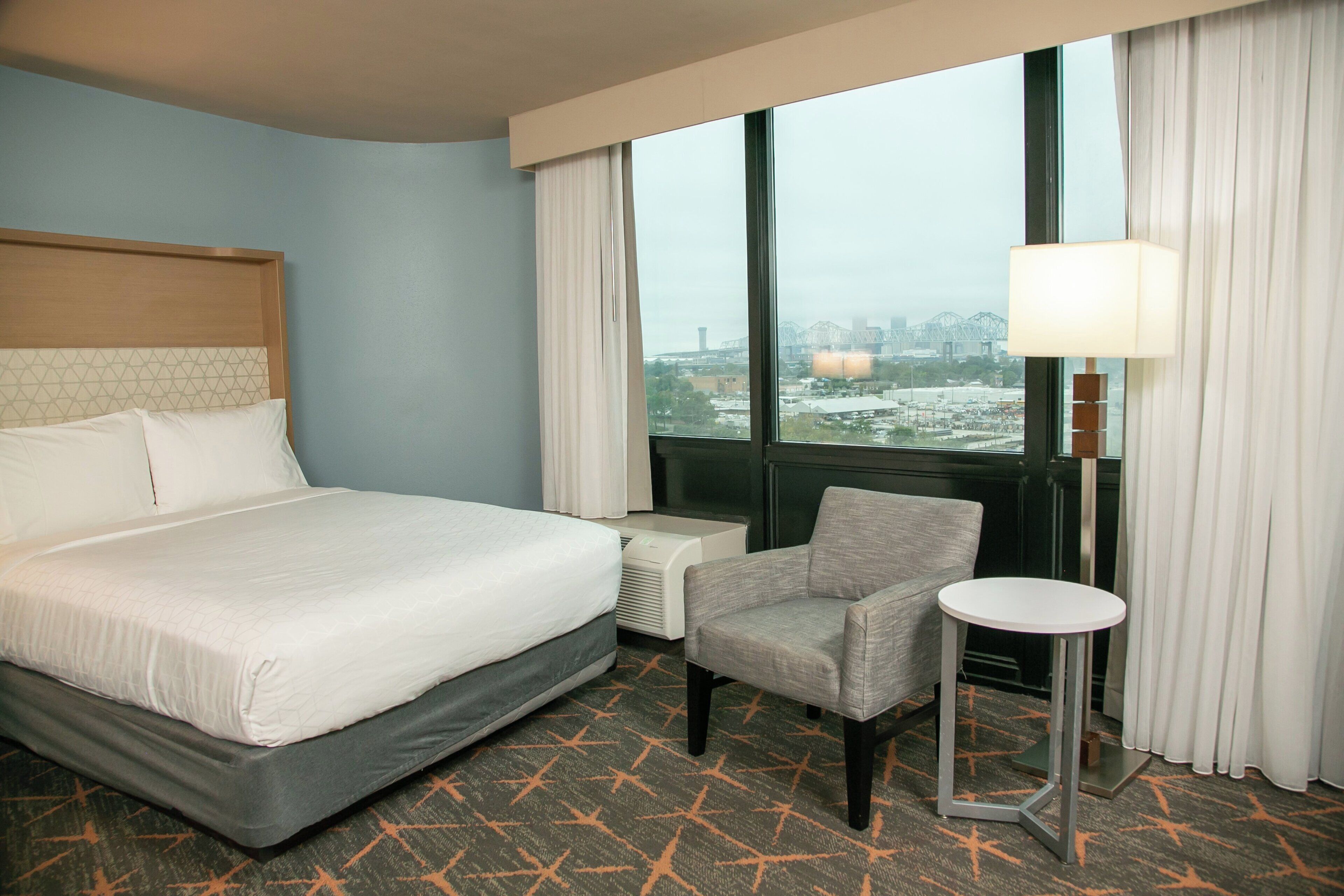 A hotel room with a bed, a chair, a small table, a window with curtains, and a city view.