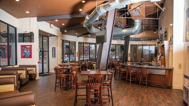 A restaurant with wooden flooring, multiple dining tables and chairs, a bar area, and large windows offering a view of the street. - Holiday Inn New Orleans West Bank Tower by IHG (Gretna)