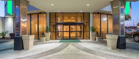 Entrada de un hotel con puerta de vidrio, plantas en macetas y un camino empedrado que lleva a la entrada.