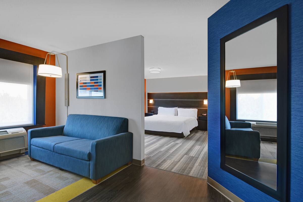 A hotel room with a blue sofa, a bed with white linens, a large mirror, and a framed picture on the wall.
