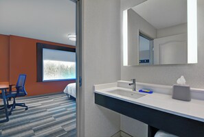 A hotel room with a bed, a desk, a chair, a bathroom with a sink and mirror, and a window with a view.