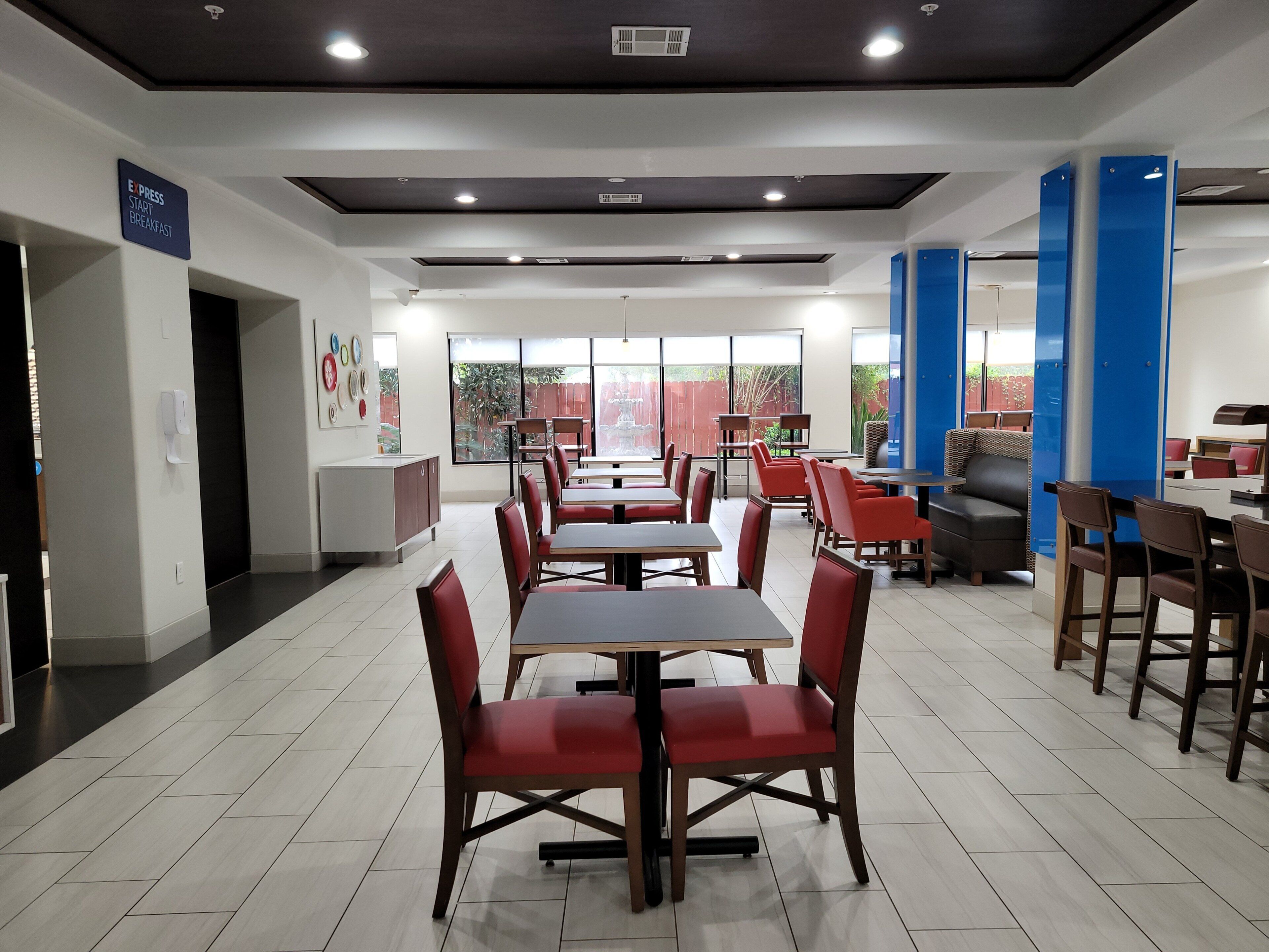 A modern dining area with tables and chairs, a sign indicating breakfast service, and a view through large windows.