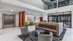 Un hall con un sofá marrón, dos sillones, una mesa redonda, un ascensor y una escalera.