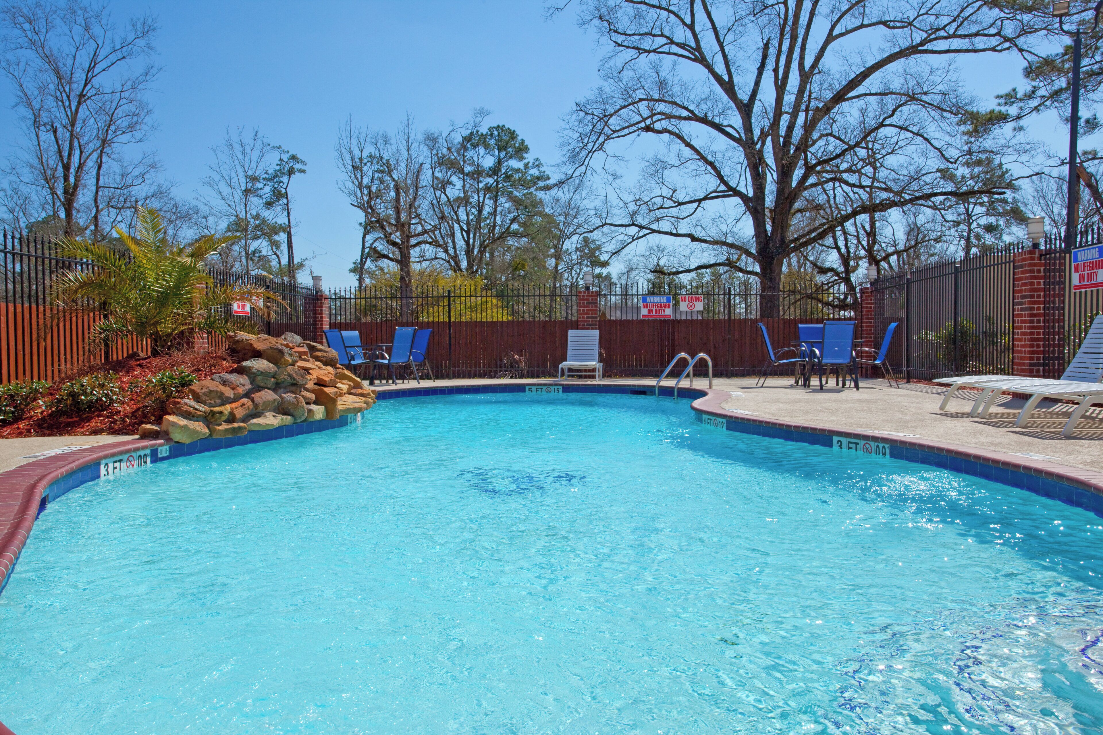 A swimming pool with a clear blue water, surrounded by a brick wall and a metal fence.