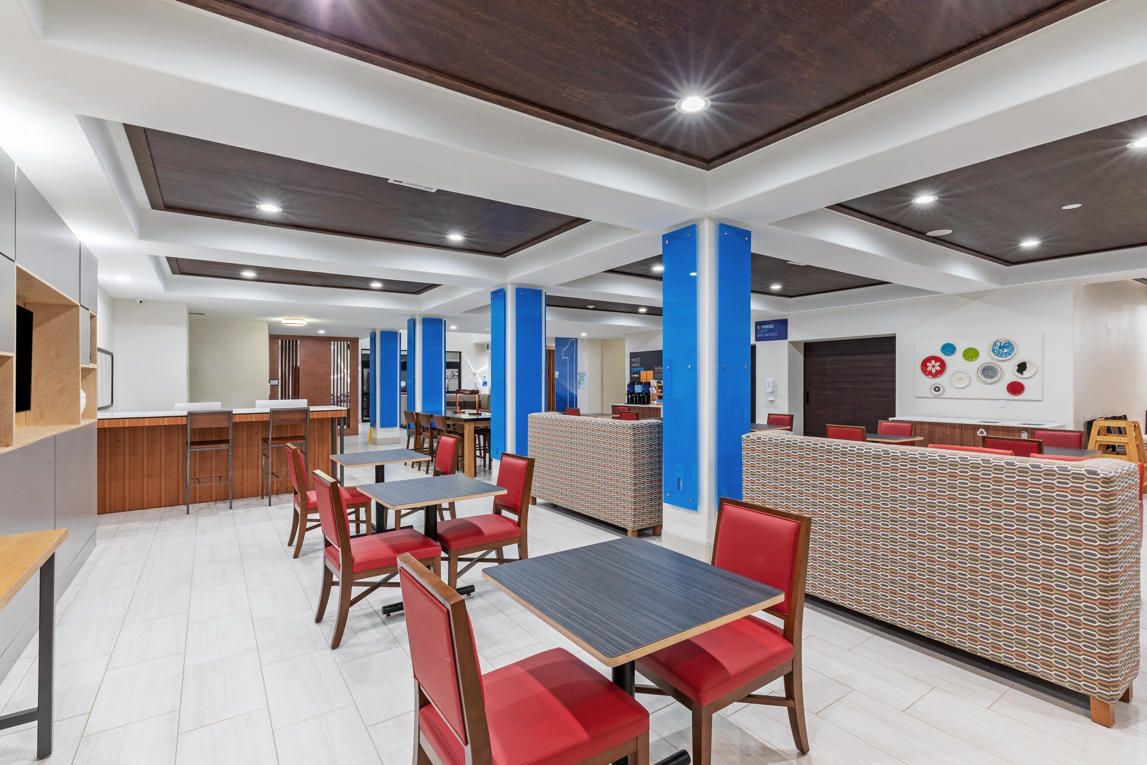 A modern cafeteria with red and wooden tables and chairs, blue pillars, and a patterned counter.