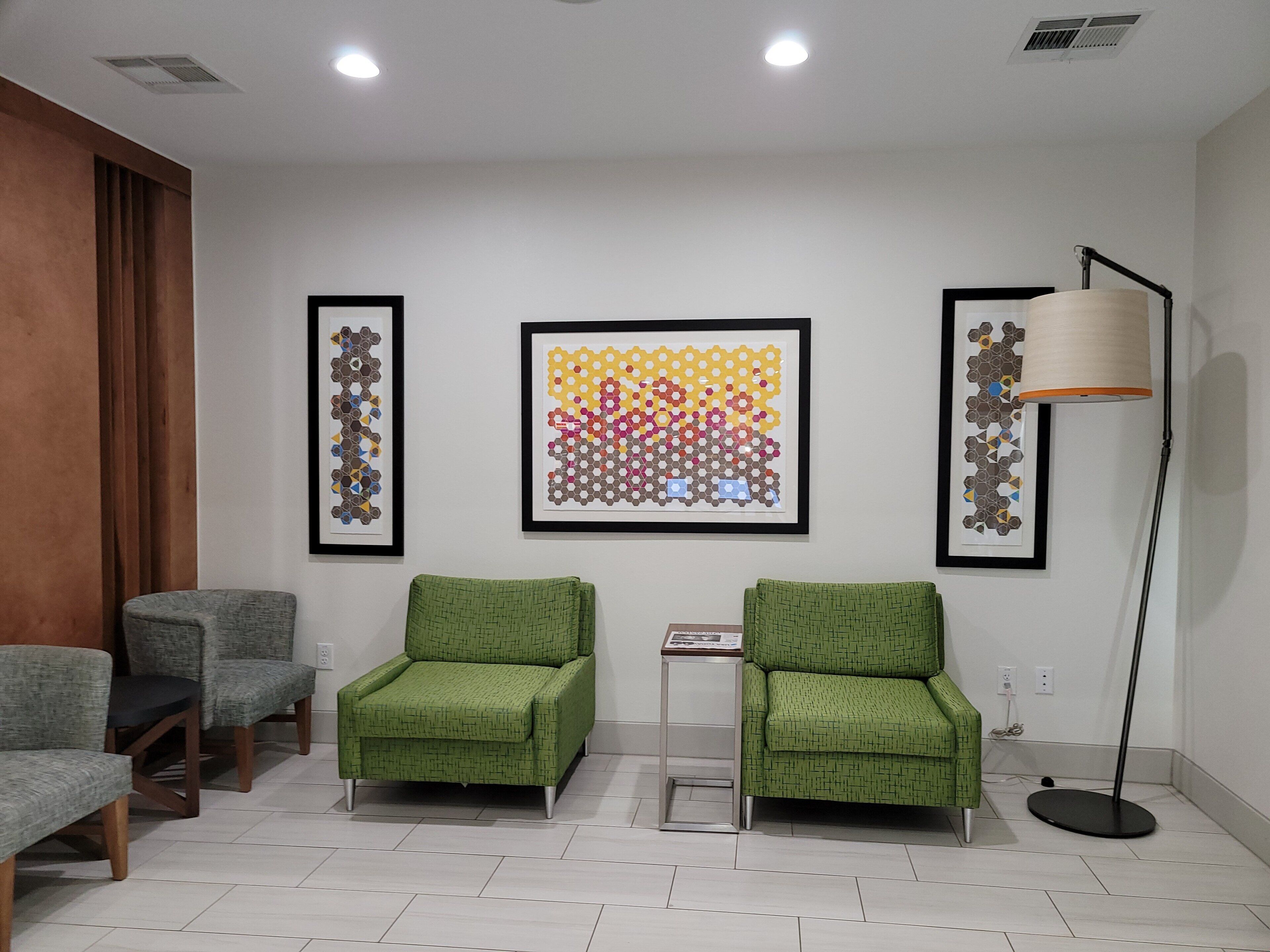 Un área de espera con dos sillones verdes, una lámpara de pie y tres obras de arte enmarcadas en la pared.