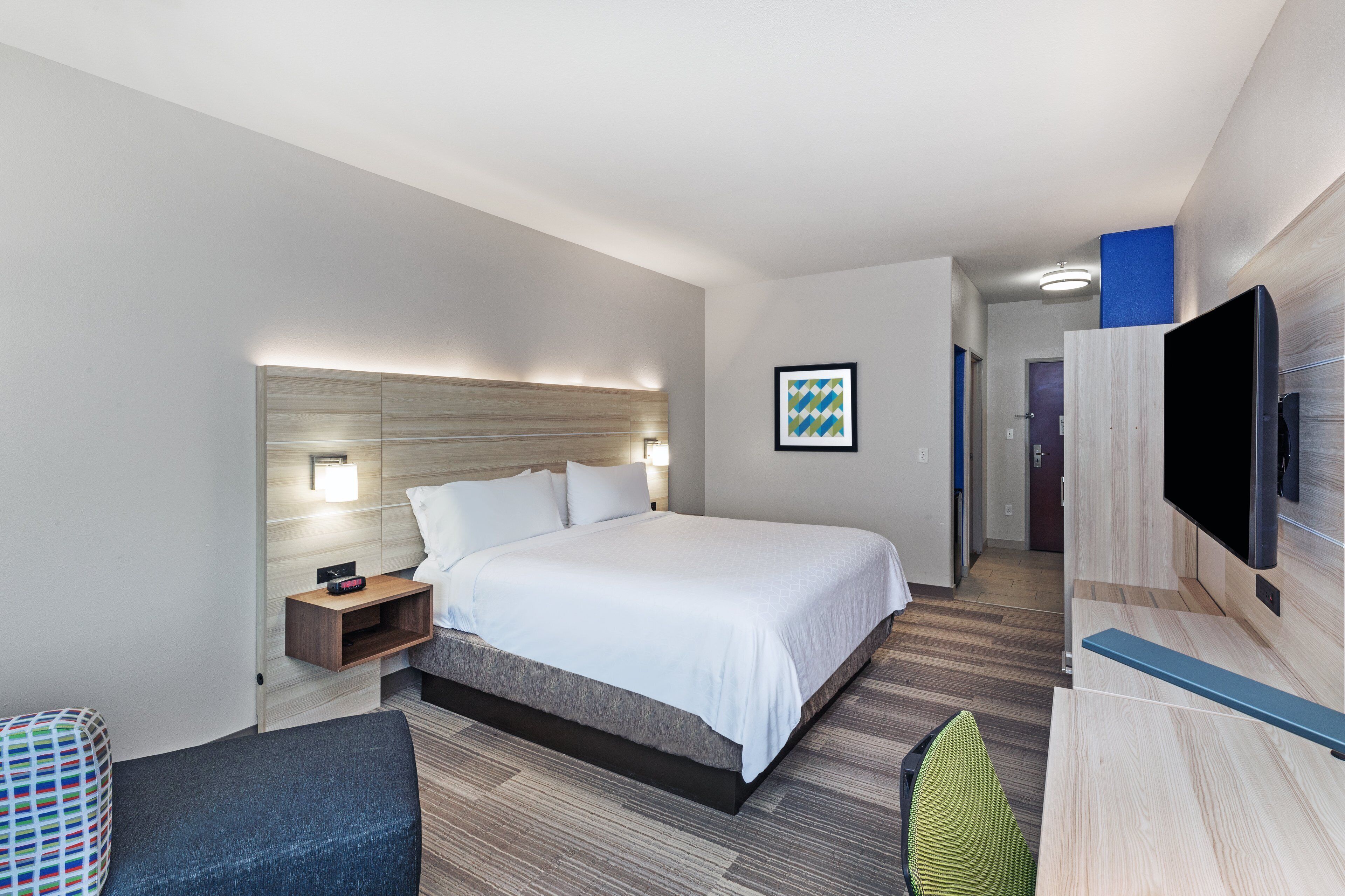 Una habitación de hotel moderna con una cama grande, mesita de noche, lámpara y un televisor de pantalla plana montado en la pared.