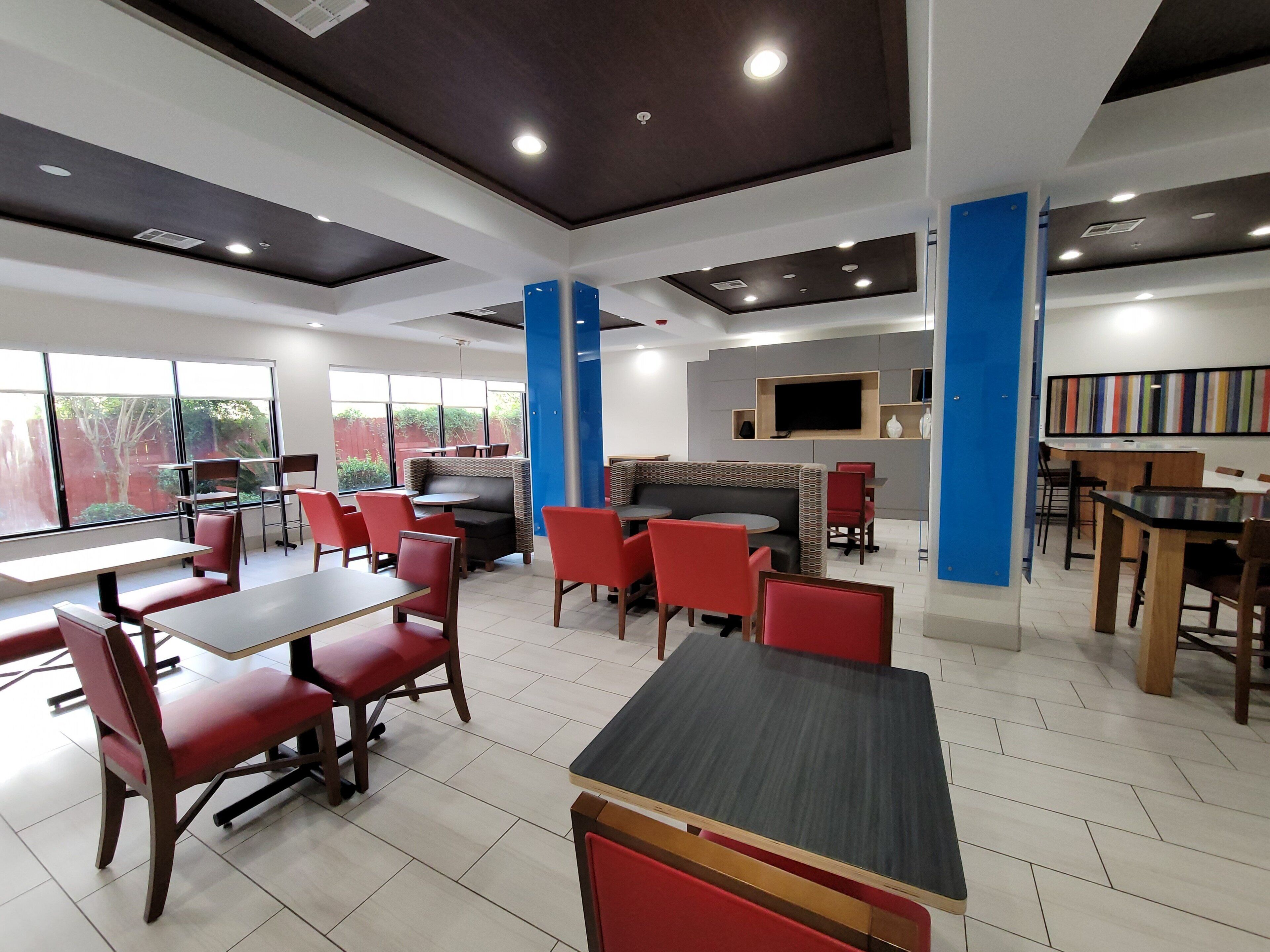 A modern restaurant interior with high tables and booths, a flat-screen TV mounted on the wall, and large windows offering a view of the outdoors.