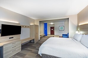 A hotel room with a bed, a TV, a desk with drawers, a bench, and a door leading to another room.