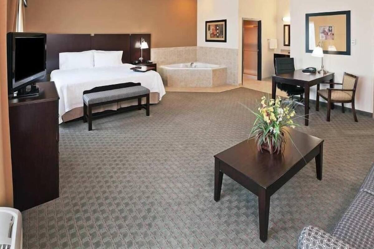 Une chambre d’hôtel avec un lit, un bureau, une chaise, une télévision, une petite table avec des fleurs, une baignoire et un tableau encadré au mur.