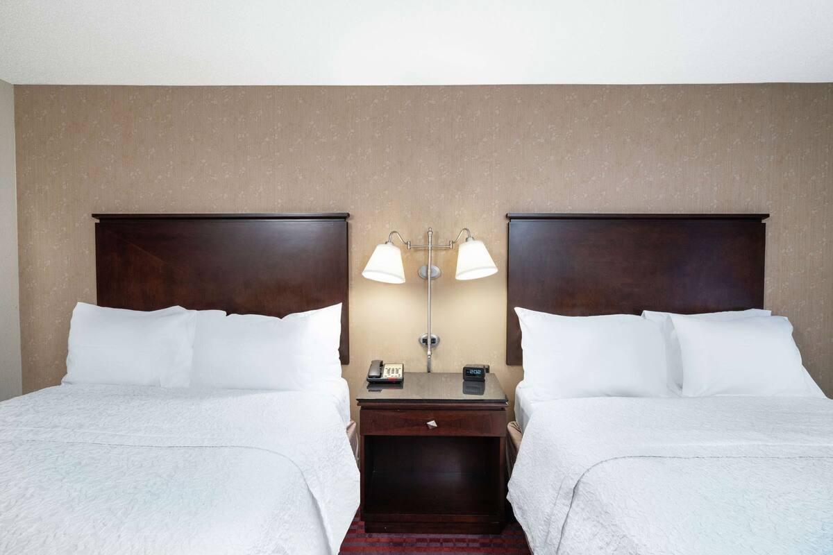 A hotel room with two beds, a nightstand with a lamp, a phone, and a digital clock.