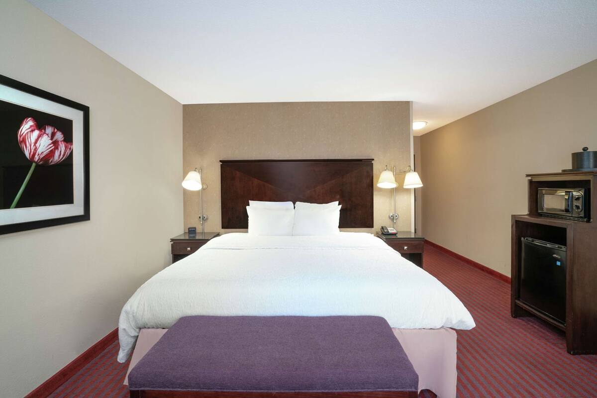 A hotel room with a large bed, bedside lamps, a small table with a lamp, a microwave, and a framed picture on the wall.