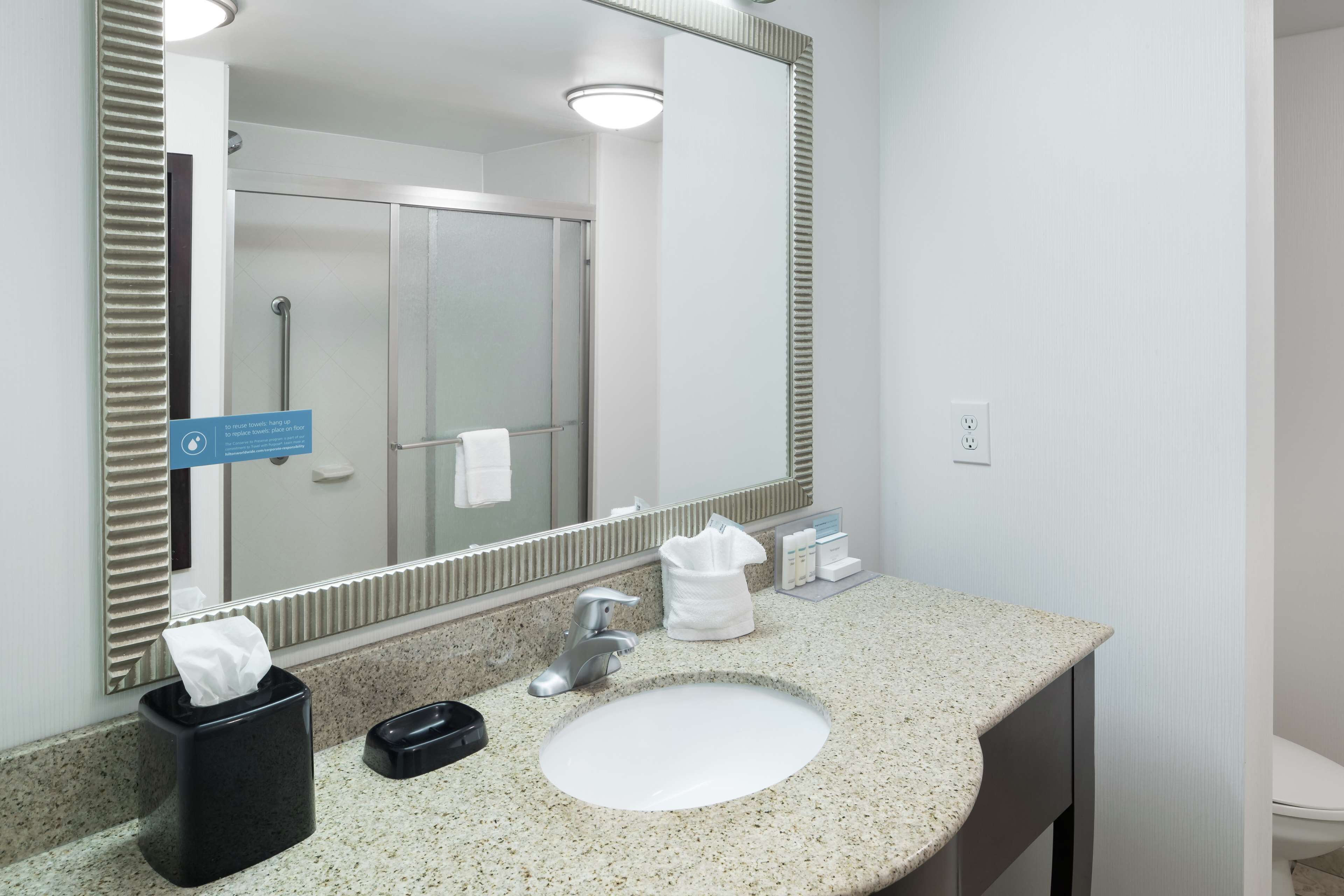 A hotel bathroom with a granite countertop sink, a mirror, a towel dispenser, and a hand dryer.
