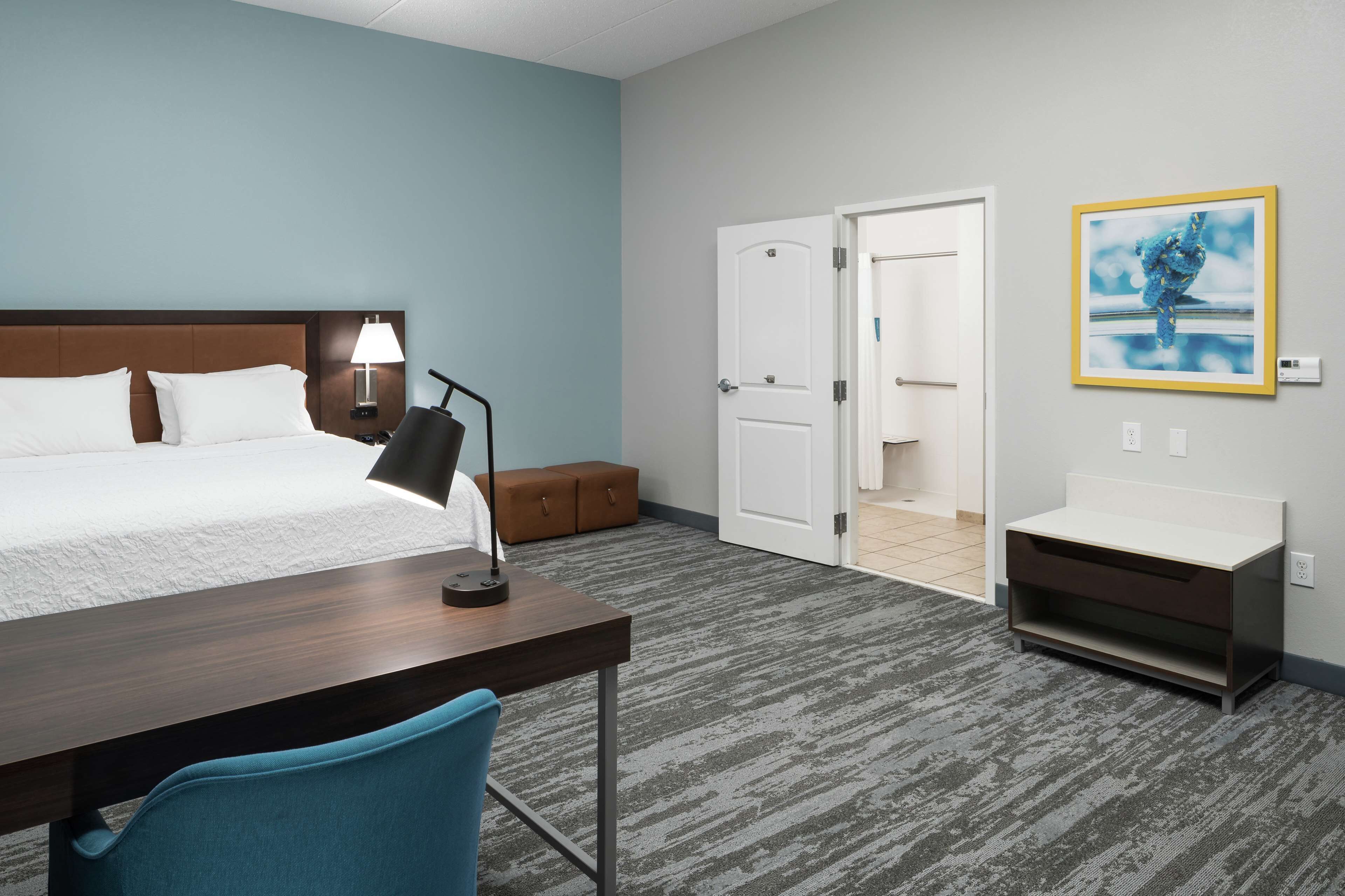 Habitación de hotel con cama, escritorio, silla, lámpara, mesita de noche y baño visible a través de una puerta abierta.