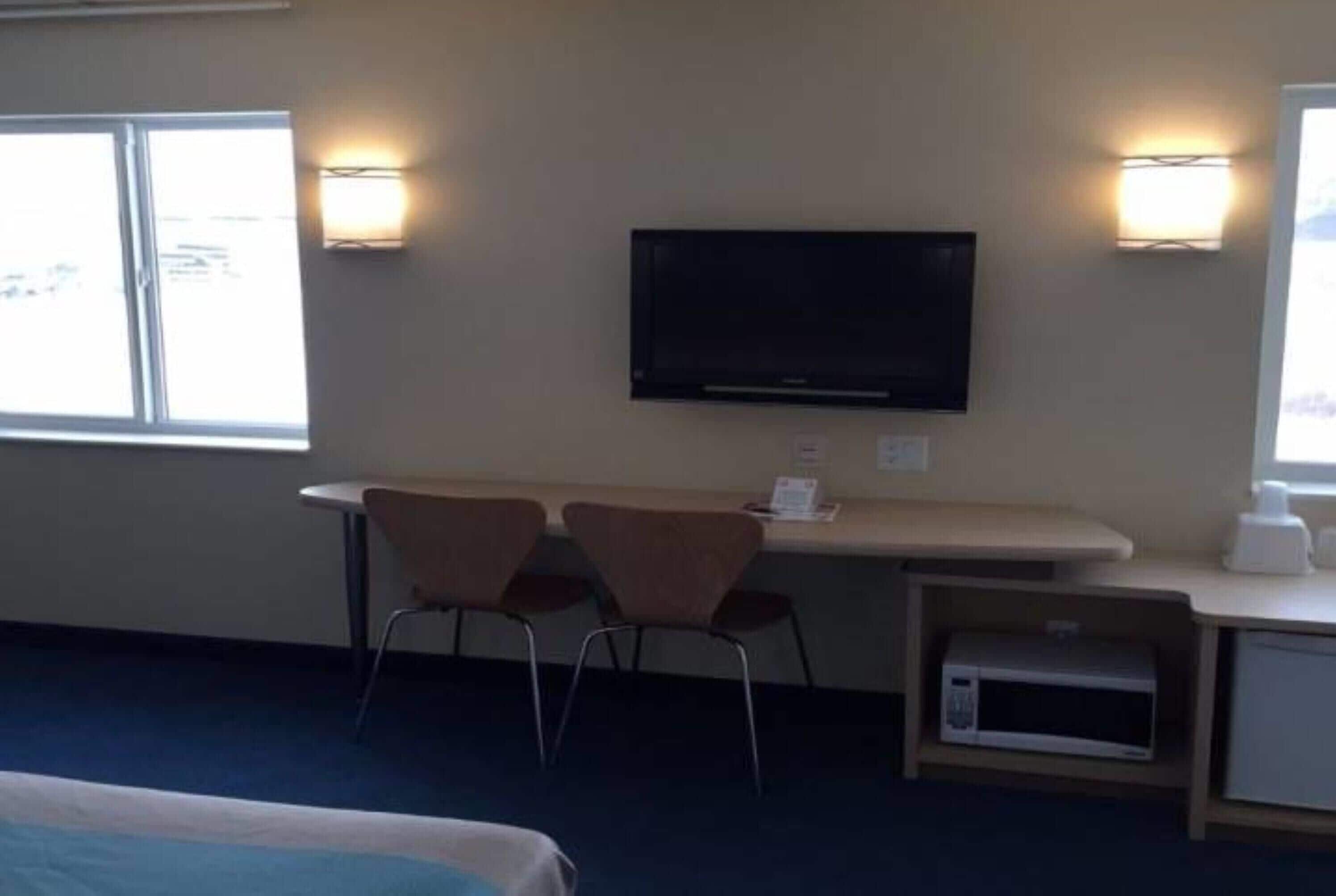 A hotel room with a bed, a desk with two chairs, a television, wall sconces, a microwave, and a window.