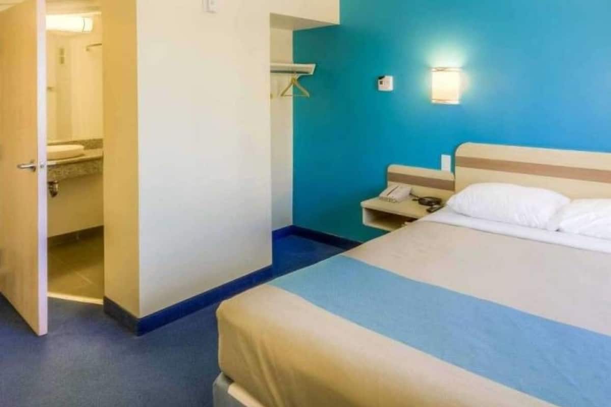 A hotel room with a bed, a nightstand, a wall-mounted light, and a door leading to a bathroom.