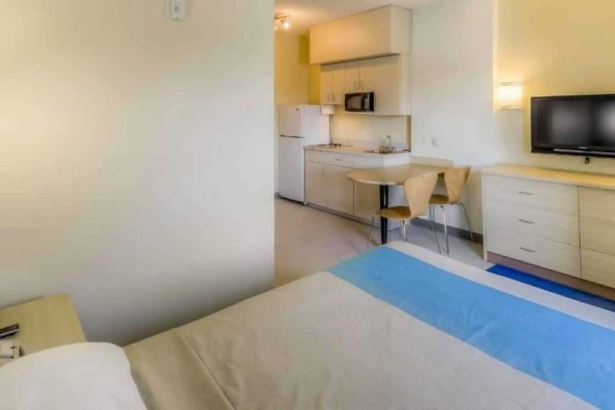A compact hotel room with a bed, a small kitchenette, a television, and a desk.