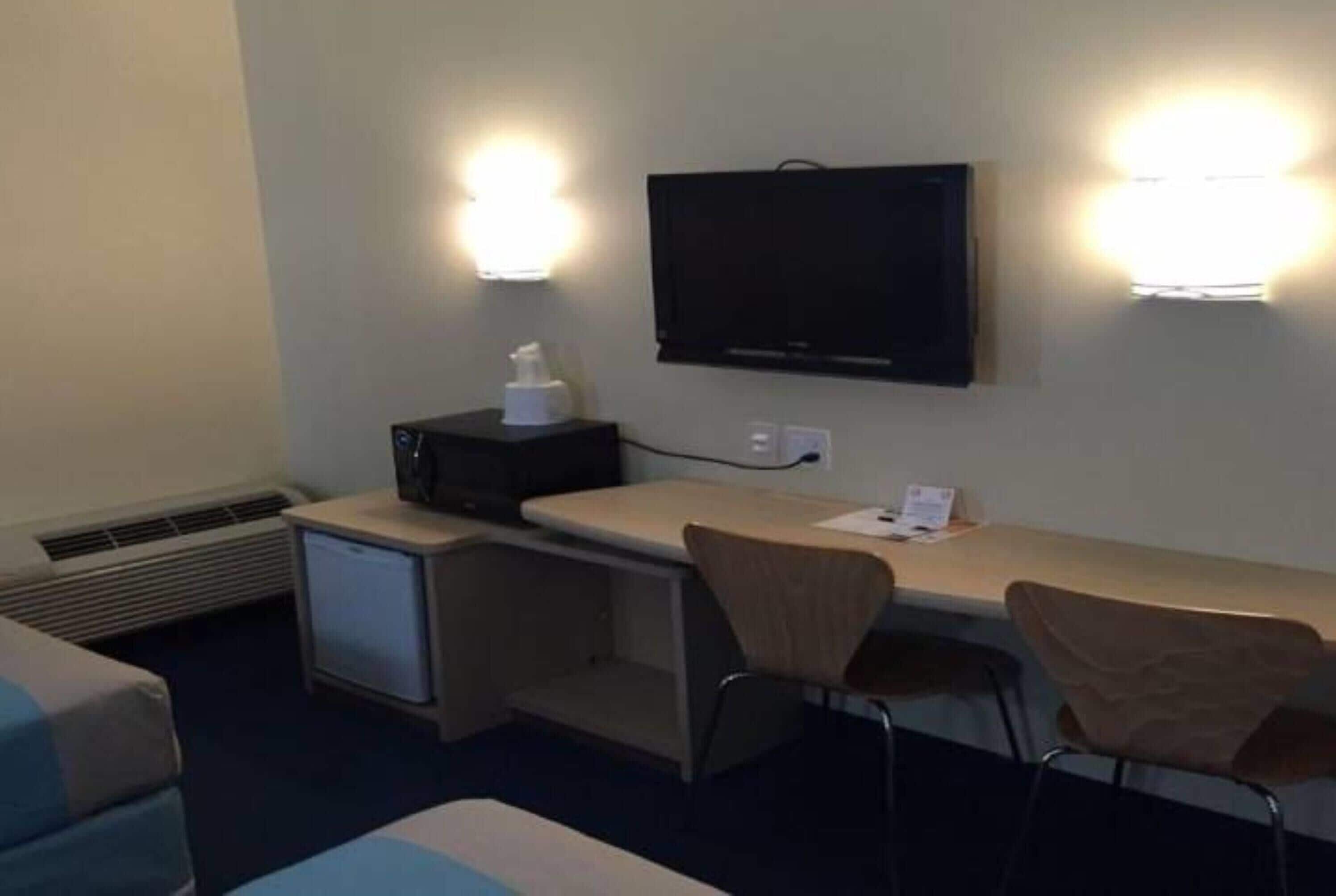 A hotel room with a bed, a desk with a chair, a television mounted on the wall, and a small heater.