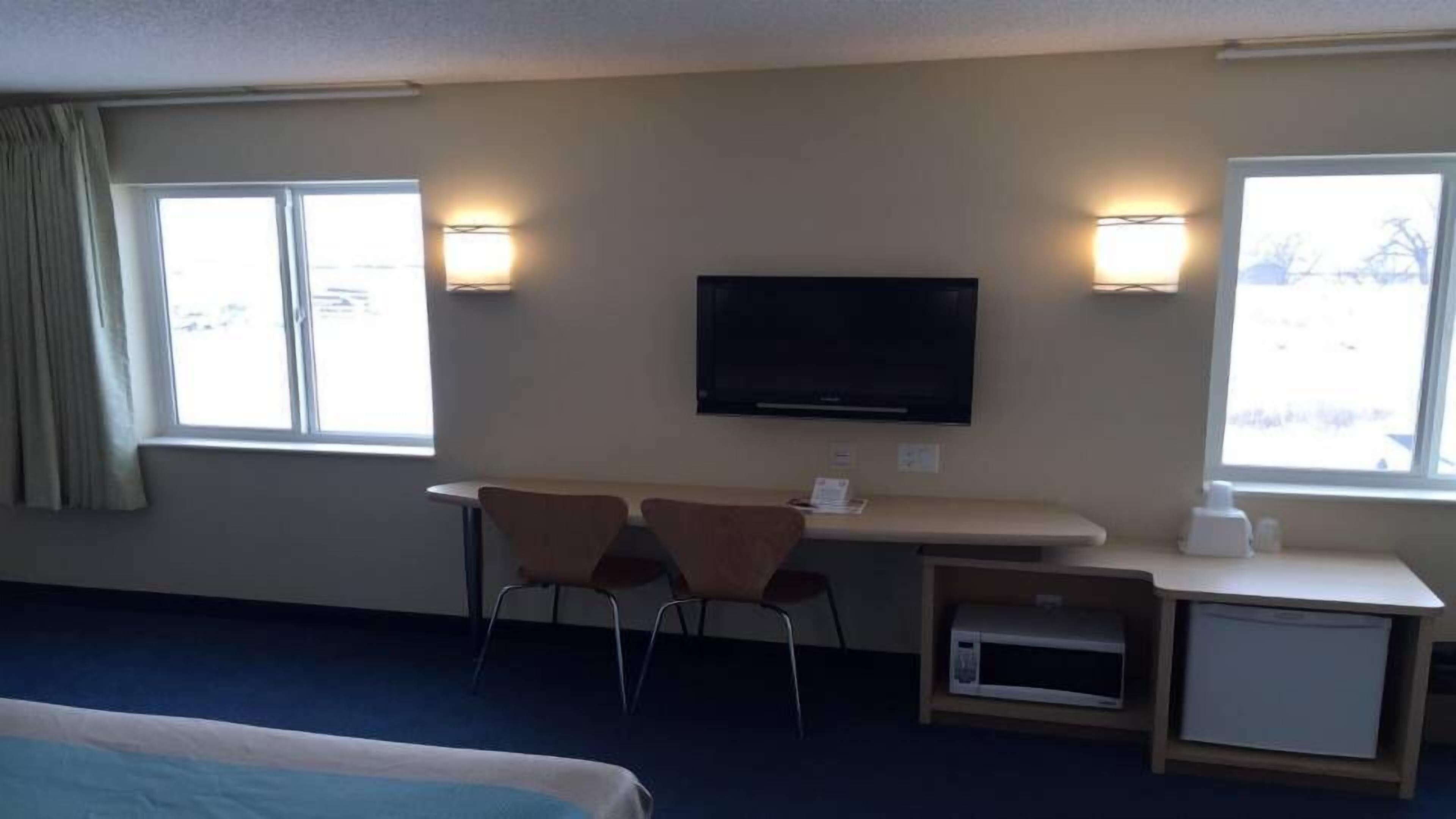 Habitación de hotel con cama, escritorio con dos sillas, televisor y dos ventanas con lámparas.