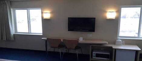 Habitación de hotel con cama, escritorio con dos sillas, televisor y dos ventanas con lámparas.