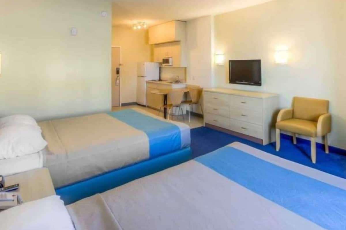 A hotel room with two beds, a desk, a chair, a television, and a kitchenette.